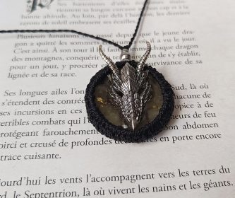 Détail du pendentif dragon entouré de tissage macramé posé sur les pages d'un grimoire ancien.