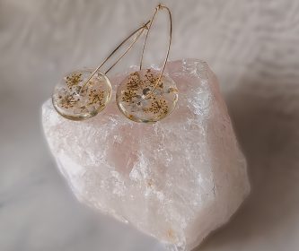 Boucles d'oreilles créoles dorées en acier inoxydable avec pendentifs donuts en résine, inclusions de fleurs séchées blanches et bleues, posées sur un quartz rose.
