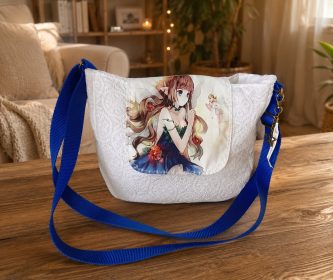 Sac bandoulière Kréa Chic en dentelle blanche avec rabat imprimé fée bleue et sangle bleue posé sur une table en bois.