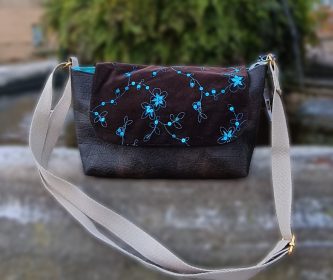 Vue de face du sac bandoulière Kréature en simili cuir marron craquelé et rabat en velours à sequins, agrémenté d'un bijou de sac artisanal en résine et d'une sangle en coton écru.